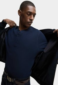 Marinblå rippad henley-tröja med tre knappar, bärs under en svart jacka. Har en figurnära design, tät väv och mörka denimjeans.