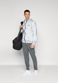 Hellgraue Zip-up-Jacke mit Kapuze, elastischen Bündchen und Logo, kombiniert mit grauen Jogginghosen mit seitlichen Streifen und weißen Sneakers.