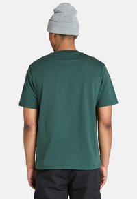 Hombre vestido con una camiseta oscura verde lisa, pantalones negros y un gorro de tejido gris claro, de pie de espaldas contra un fondo blanco.