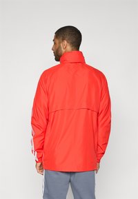 Muž oblečený v červenej bunde s bielymi pruhmi na rukávoch a sivými nohavicami, zobrazený zozadu na jednoduchom svetlom pozadí.