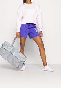 Světle růžová krátká mikina, fialové šortky s textem "just do it" a šedá sportovní taška. Bílé boty s jemnými oranžovými akcenty.