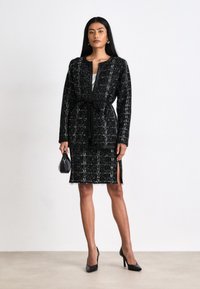 Robe en tweed noir et blanc avec une taille ceinturée, des manches longues et une jupe dotée de fentes latérales. Associée à des escarpins noirs et un sac à main.