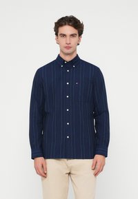 Tengerészkék, függőleges fehér és piros csíkos ing, mellzsebes és galléros kivitelben, világos bézs nadrággal viselve.