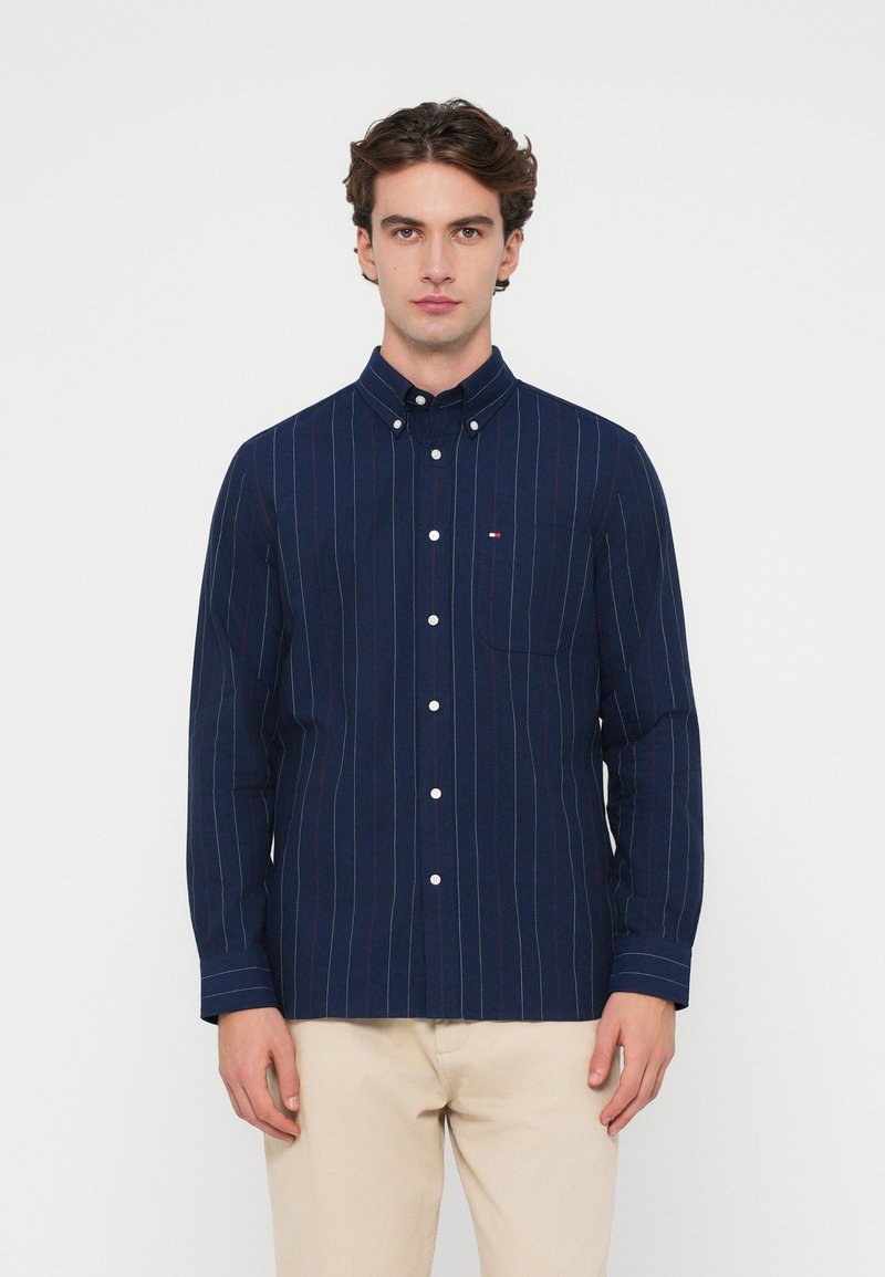 Tengerészkék, függőleges fehér és piros csíkos ing, mellzsebes és galléros kivitelben, világos bézs nadrággal viselve.