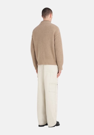 Pull en maille beige avec un col montant et une texture côtelée, associé à un pantalon crème à coupe décontractée avec poches latérales. Des chaussures noires terminent le look.