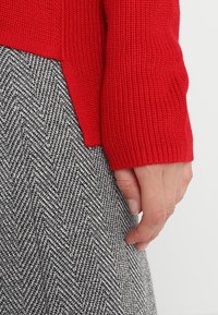 Pull en tricot rouge avec un tissu texturé, associé à une jupe à motif pied-de-poule noir et blanc. Main visible reposant sur le bord de la jupe.