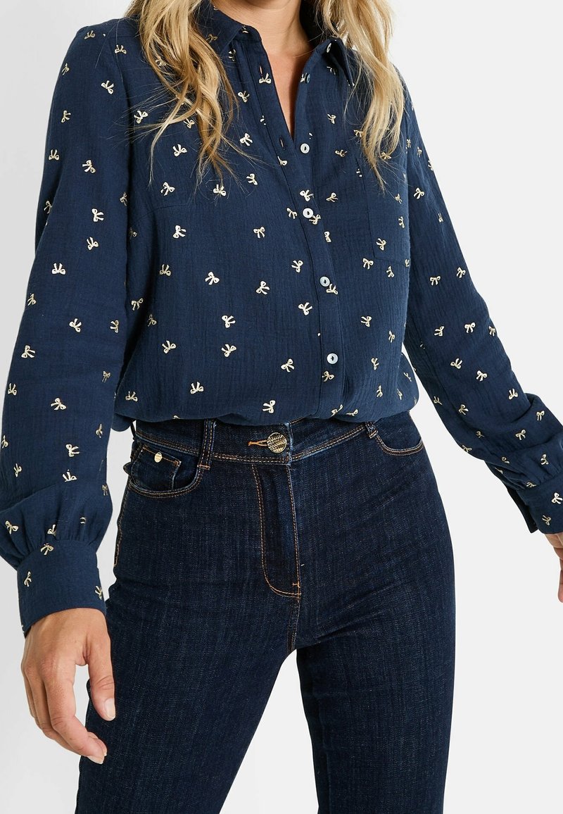 Femme portant un chemisier bleu marine boutonné avec de petits motifs de nœuds blancs et un jean bleu foncé taille haute, debout devant un fond uni.