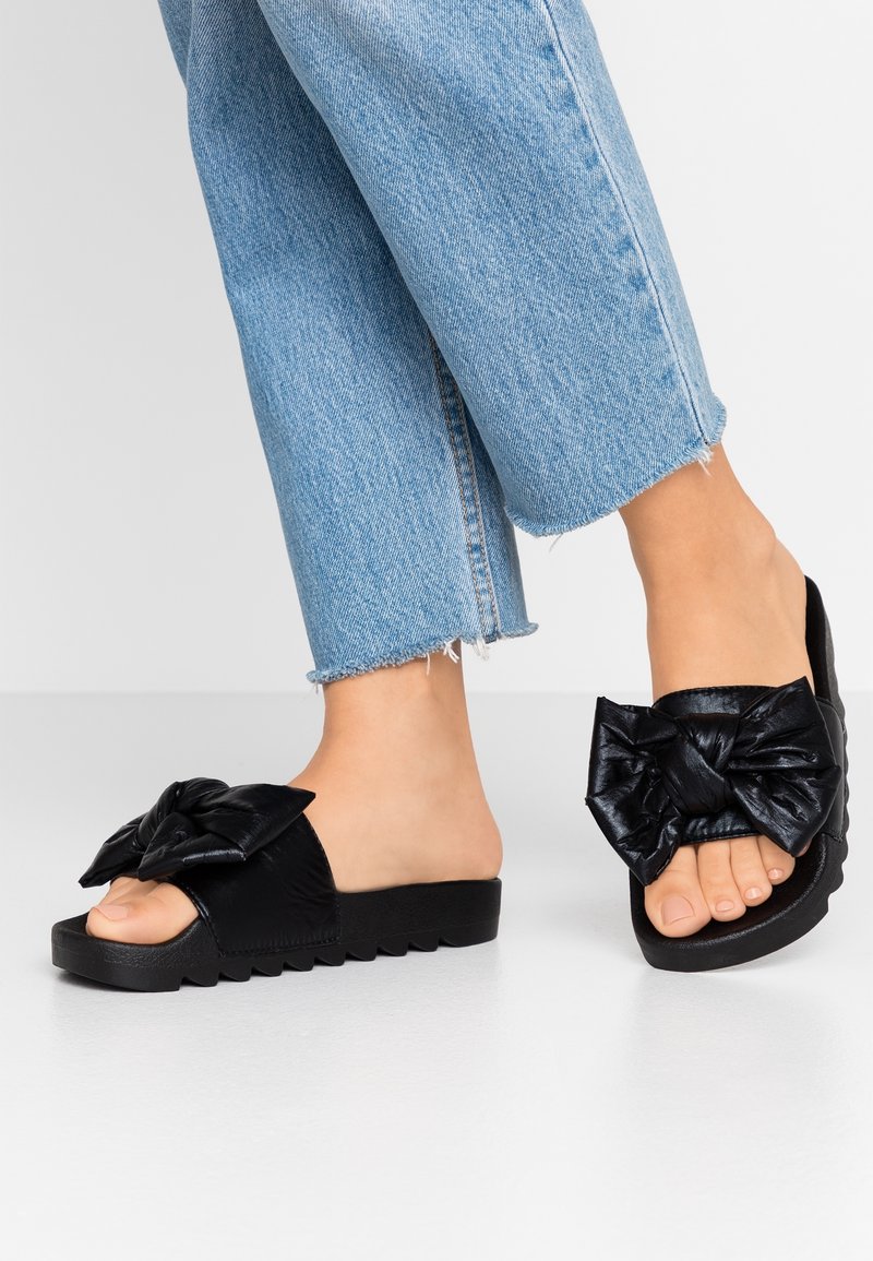 Feet wearing black slide sandals with large bows paired with light blue frayed hem jeans on white floor.