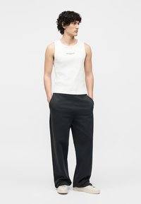 Άνδρας με σγουρά μαλλιά που φοράει λευκό αμάνικο τοπ, άνετο μαύρο παντελόνι και λευκά αθλητικά παπούτσια, στέκεται με τα χέρια στις τσέπες και κοιτάζει δεξιά.