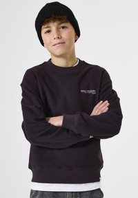 Sudadera de color púrpura oscuro con puños y dobladillo acanalados, que presenta un pequeño logo "TOTAL SOLITUDE" en púrpura claro. Lleva puesta una camisa blanca debajo.