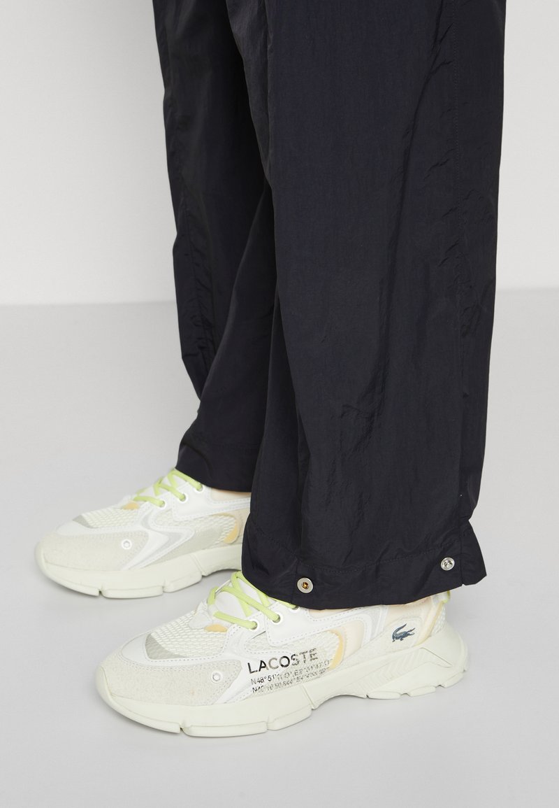 Witte sportieve sneakers met een textuur van mesh en suède accenten, voorzien van neongroene veters en een merklogo aan de zijkant. Gecombineerd met zwarte broek.