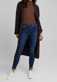 Suéter marrón de punto, cárdigan largo negro y con manchas multicolores, jeans ajustados oscuros y zapatillas blancas.