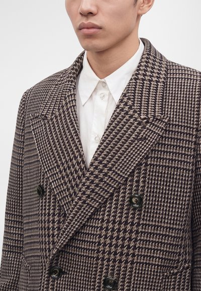 Homme portant un manteau croisé marron et beige à motif pied-de-poule sur une chemise blanche boutonnée, montré du cou jusqu'au milieu du torse.