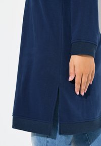 Tunic blu navy con taglio dritto, polsini a coste e spacco laterale. Il materiale sembra morbido e leggermente testurizzato. Mano appoggiata sul tessuto.