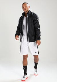 Svart lett jakke med hvit glidelås og logo, brukt over en hvit skjorte og shorts, complementert med svarte sokker og hvite/røde sko.