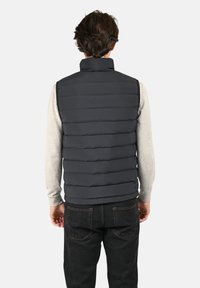 Gilet matelassé gris foncé avec col haut, présentant des sections matelassées horizontales, porté sur une chemise à manches longues gris clair et un jean foncé.