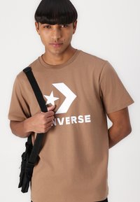 Ruskea puuvillapaita, jossa on suuri valkoinen "CONVERSE"-logo, tähti ja nuolimuotoilu. Malli pitää mustaa olkalaukkua olkapäällään.