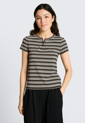 Junge Frau mit dunklen Haaren, die ein schwarz-weiß gestreiftes kurzärmeliges Oberteil und schwarze Hose trägt, steht mit einer Hand in der Tasche.