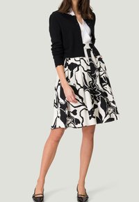 Zwarte cardigan over een witte blouse met kraag, gecombineerd met een geplooide zwart-witte bloemenrok. Zwarte schoenen maken de outfit compleet.