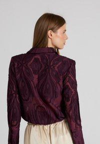 Paarse geborduurde blouse met ingewikkelde bloemenmotieven, lange mouwen en een kraag, gemaakt van een gestructureerde, transparante stof.