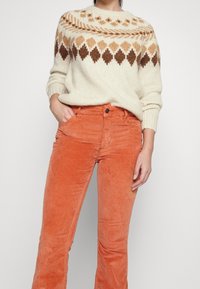 Pull crème en tricot avec motif losanges marron et beige, associé à un pantalon en velours côtelé orange. Le pull présente un col rond et des poignets côtelés.