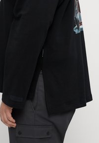 Black oversized long-sleeve shirt with a curved hem, side slits, and a graphic print on the sleeve. Soft cotton fabric.
