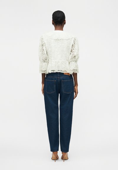 Femme avec les cheveux en chignon portant un chemisier blanc en dentelle avec des manches bouffantes, un jean bleu taille haute et des sandales beiges à talons hauts, debout.