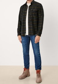 Camisa de franela a cuadros en verde oscuro y negro, camiseta de manga larga color beige debajo, jeans azules y botas beige con cordones naranjas.