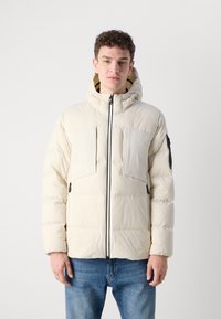 Jachetă pufoasă de culoare bej deschis, cu glugă, fermoar frontal negru, două mari buzunare la piept și mansete ribbate. Material neted, izolat.