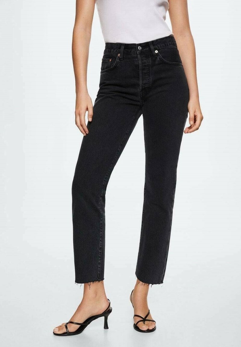Femme portant un jean noir taille haute, coupe droite, avec des ourlets bruts et des sandales noires à talons avec brides, associé à un haut blanc sans manches.