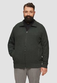 Dunkelgrüner Zip-Up-Cardigan mit einem strukturierten Strickmuster, seitlichen Taschen und gerippten Manschetten und Saum. Über einem schwarzen Shirt getragen.