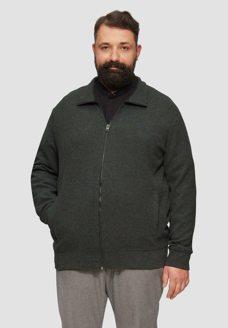 Dunkelgrüner Zip-Up-Cardigan mit einem strukturierten Strickmuster, seitlichen Taschen und gerippten Manschetten und Saum. Über einem schwarzen Shirt getragen.