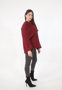 Top rosso sovradimensionato con texture e colletto drappeggiato unico, abbinato a jeans grigi aderenti e stivaletti neri con punte appuntite.