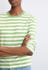 Groen en wit gestreepte lange mouwen T-shirt met een ronde hals en geribde manchetten, gedragen met gekruiste armen. Zachte katoenstof.