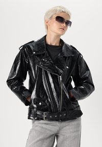 Juodas dirbtinės odos motociklininkų švarkas su blizgiu paviršiumi, užtrauktuku, dirželiu juosmens srityje ir sidabrinėmis detalėmis. Dėvimas virš juodos marškinės.