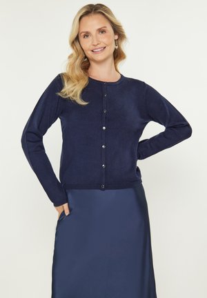 Navy cardigan met lange mouwen, een ronde halslijn en knoopsluiting aan de voorkant. Zachte textuur, slim fit ontwerp, gecombineerd met een satijnen marineblauwe rok.