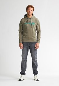 Felpa verde oliva con testo stampato teal, cordini e polsini a coste, abbinata a jeans grigi e sneakers bianche. Design minimale.