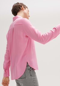 Personne portant une chemise rose ample à manches longues avec un ourlet incurvé et un pantalon gris, debout devant un fond clair uni.