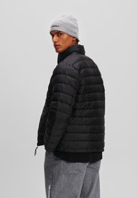 Zwarte pufferjack met gequilte ontwerp, hoge kraag en ritssluiting. Draag met een lichtgrijze beanie. Achtergrond is eenvoudig.