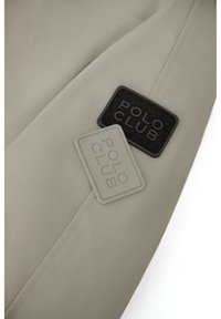 Chaqueta gris claro que presenta tres parches de goma "POLO CLUB" en negro y gris, con una textura lisa y mate y un diseño minimalista.