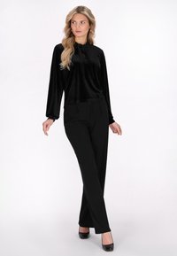 Top lungo in velluto nero a maniche lunghe con dettaglio al collo a fiocco, abbinato a pantaloni neri su misura e tacchi chiusi neri su uno sfondo chiaro.