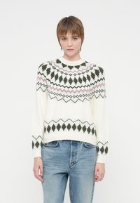 Mlad človek s kratkimi lasmi, oblečen v belo pleteno pulover z črnimi in roza geometrijskimi vzorci ter svetlo modre kavbojke, ki stoji pred belo ozadjem.