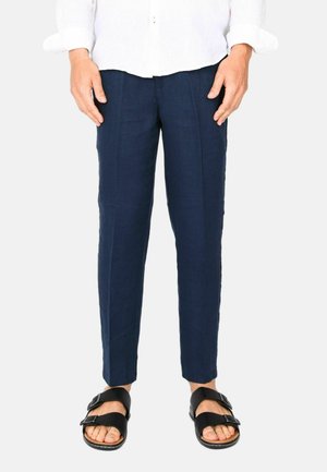 Uomo che indossa pantaloni su misura blu navy, sandali neri con doppio cinturino e una camicia bianca a maniche lunghe, in piedi contro uno sfondo semplice.