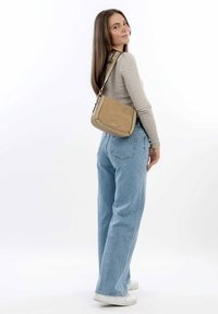 Torebka crossbody w kolorze tan, o gładkiej teksturze, zaokrąglonym kształcie i subtelnych detalach szycia. Nosi ją w zestawieniu z jasnoniebieskimi szerokimi spodniami dżinsowymi i białymi trampkami.