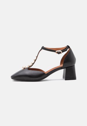 Chaussure en cuir noir à talon moyen avec bride en T, clous ronds dorés sur le devant, et fermeture à boucle latérale sur fond blanc uni.