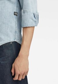 Light blue denim shirt with rolled sleeves, featuring a black logo patch on the side. Paired with dark denim jeans, showing a fitted design.