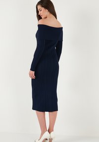Abito aderente a coste navy con spalle scoperte, maniche lunghe, lunghezza midi, tessuto testurizzato e leggero elastico; abbinato a tacchi bianchi.