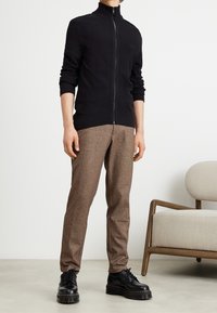 Svart zip-up sweater med hög krage, ribbade ärmslut och nederkant; i kombination med bruna strukturerade byxor och svarta snörskor.