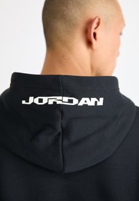 Čierna mikina s kapucňou s jemnou textúrou, podšívkovanou kapucňou a bielym nápisom "JORDAN" na chrbte blízko výstrihu.