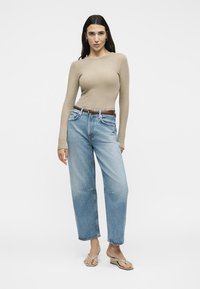 Γυναίκα στέκεται φορώντας ένα μπεζ ριμπ πλεκτό τοπ με μακριά μανίκια, ανοιχτό μπλε ψηλόμεσο τζιν, καφέ ζώνη και μπεζ πέδιλα με τακούνι.